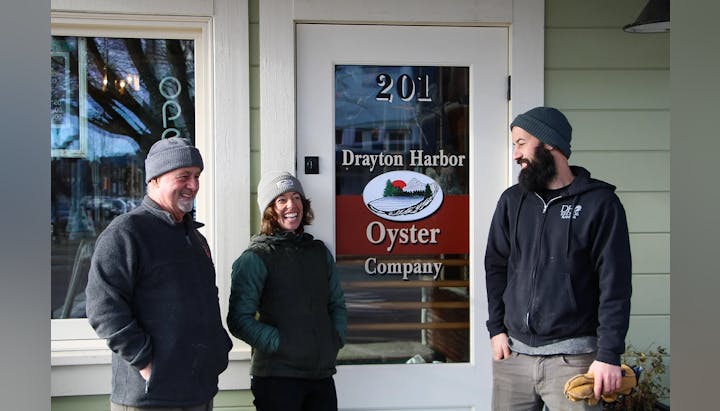 Three people in beanies smiling in front of a door marked "Drayton Harbor Oyster Company."