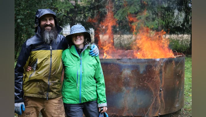 Two people in rain gear stand smiling in front of a large, blazing fire pit outdoors.