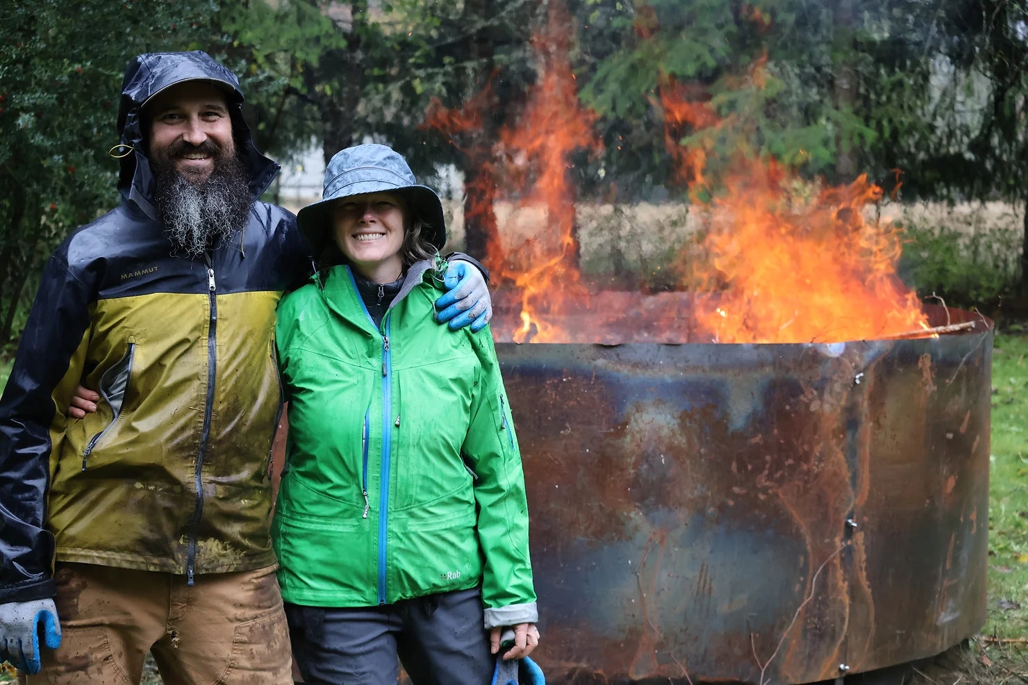 Two people in rain gear stand smiling in front of a large, blazing fire pit outdoors.