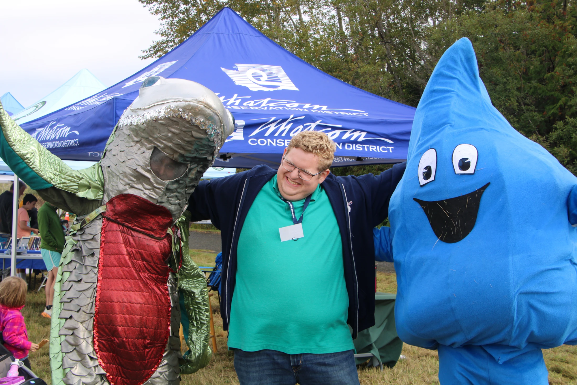 A person posing with someone in a fish costume and a blue water droplet mascot at an outdoor event.