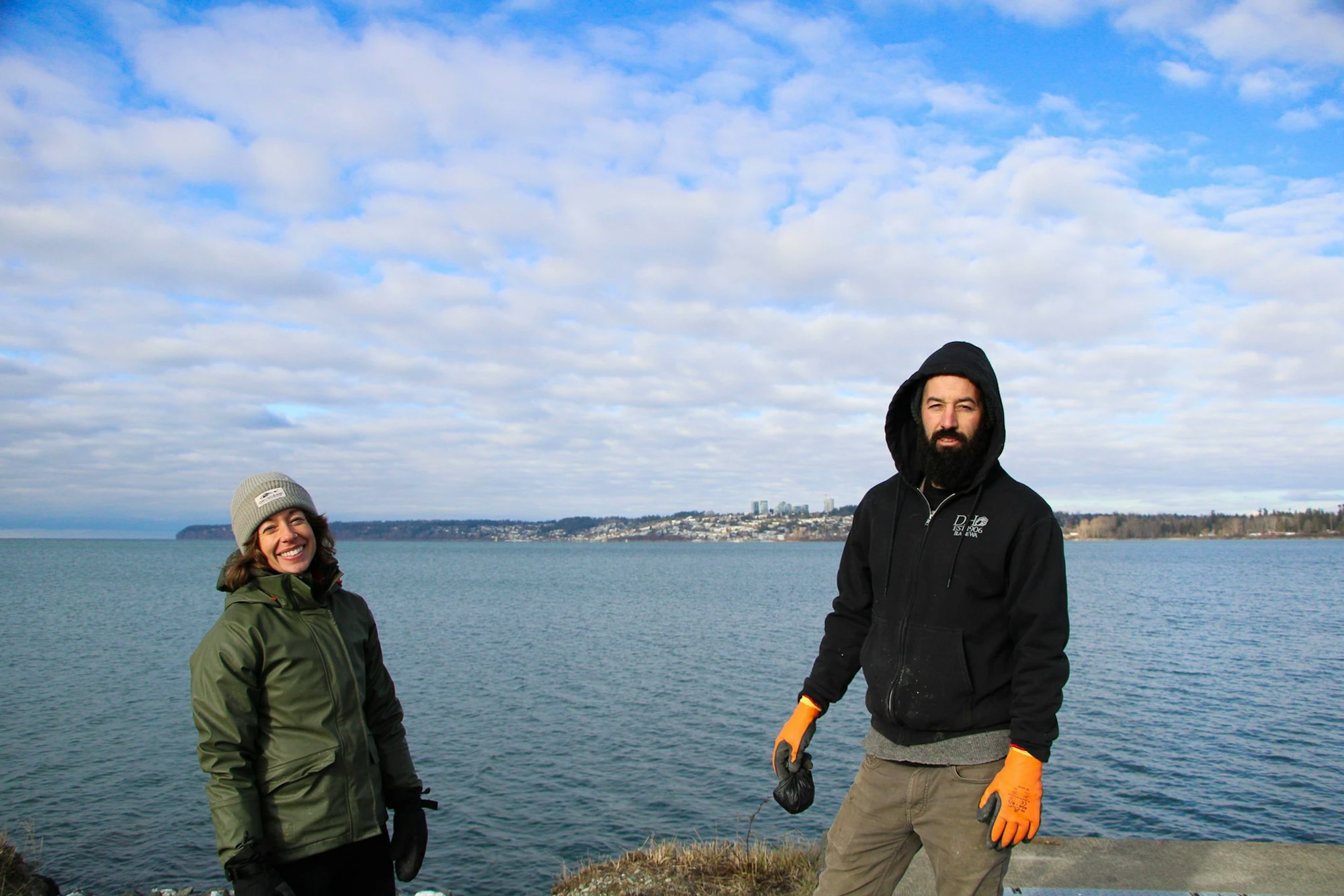 Two people in jackets and hats standing by a body of water with a cityscape in the background.