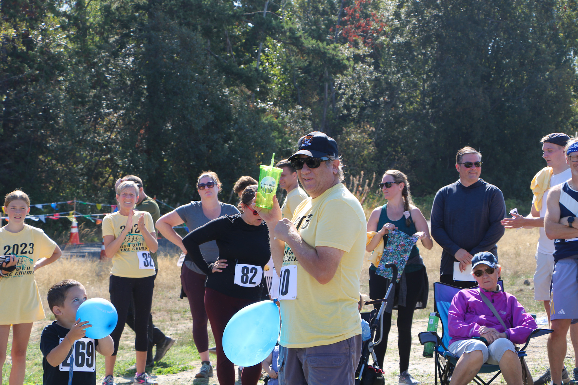 A group of people at an outdoor event, some with numbered bibs, holding balloons and drinks.