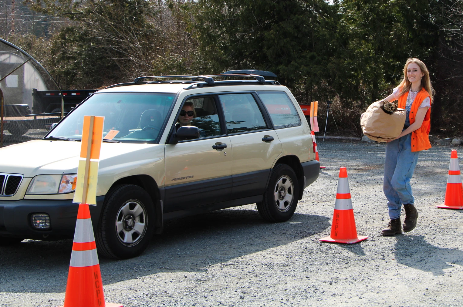 A person in a car and another carrying a paper bag while wearing an orange vest, surrounded by traffic cones in an outdoor setting.