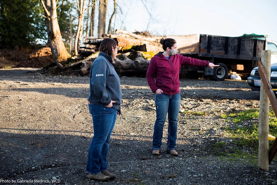 Two people outdoors; one points, the other watches. Truck, logs, and trees in the background.