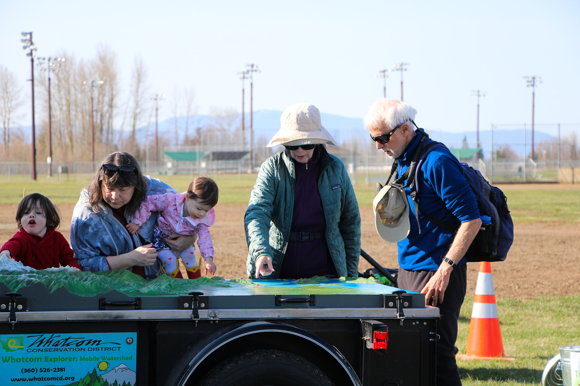 A group of people, including children, are gathered around a mobile watershed exhibit outdoors.