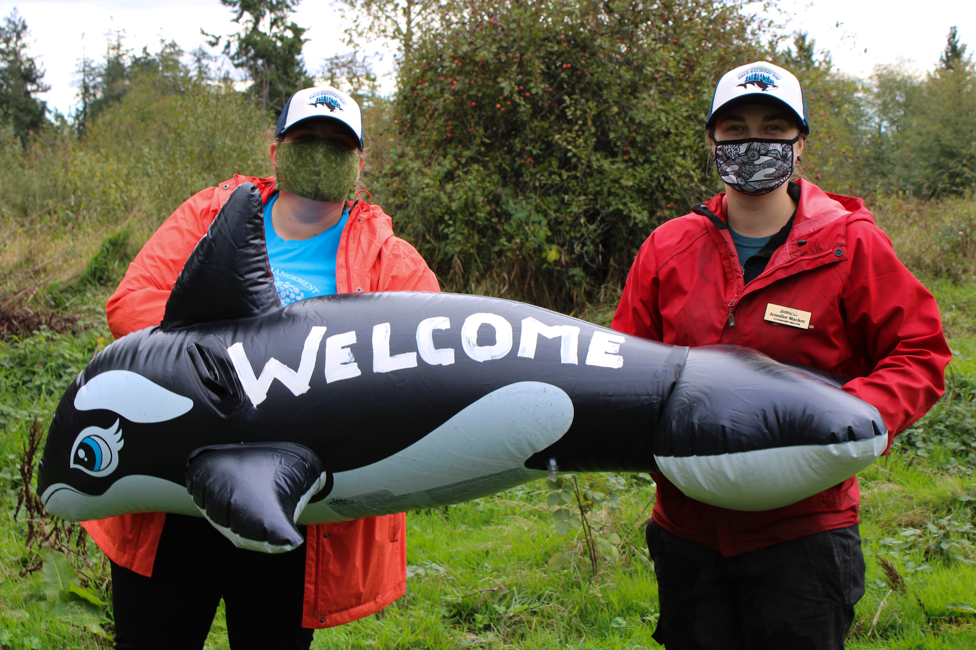 Two people in red jackets, wearing masks, holding an inflatable orca with "WELCOME" written on it.