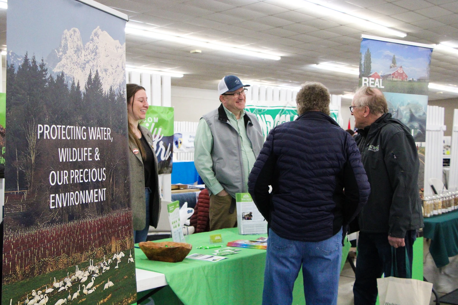 People at an environmental booth discuss conservation. A banner reads: "Protecting Water, Wildlife & Our Precious Environment."