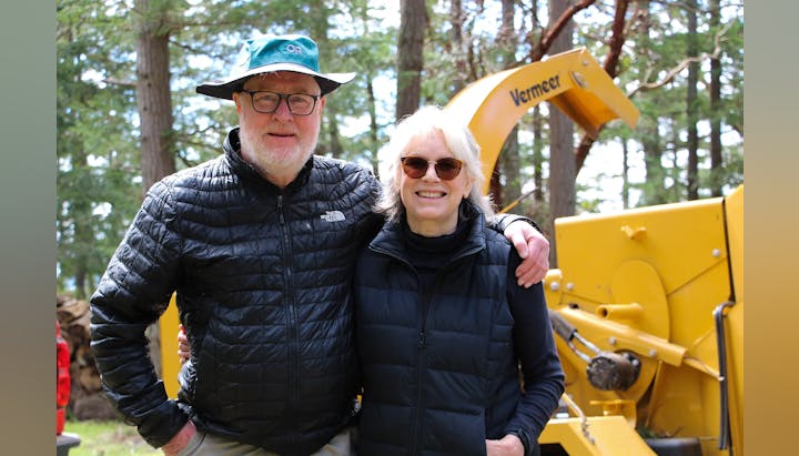 A couple in black jackets smiling outdoors, next to a yellow machine, surrounded by trees.