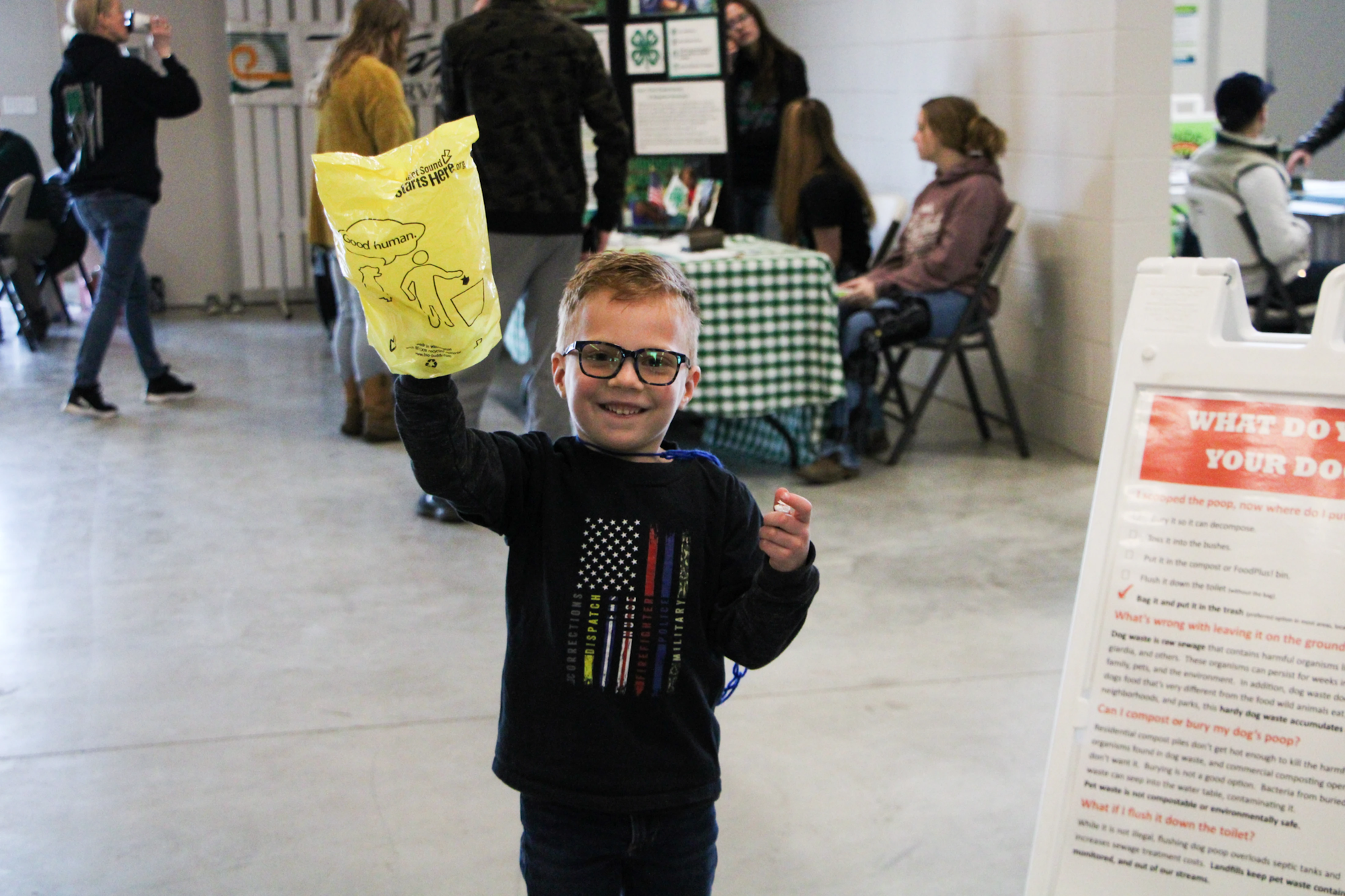 A smiling child holds a yellow bag with “Good human” on it, in a room with people and an informational sign nearby.