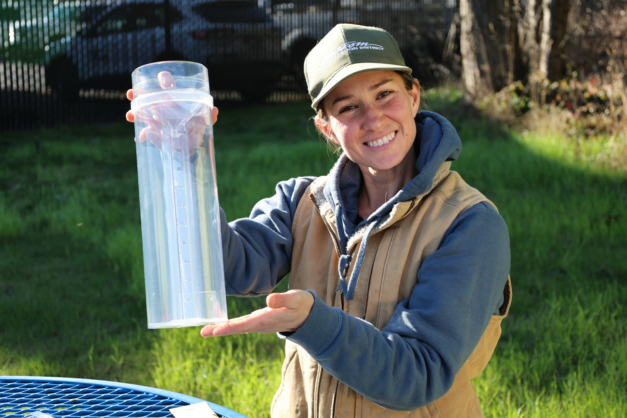 A smiling person holds a clear measuring cylinder outdoors, showcasing its scale, with green grass in the background.
