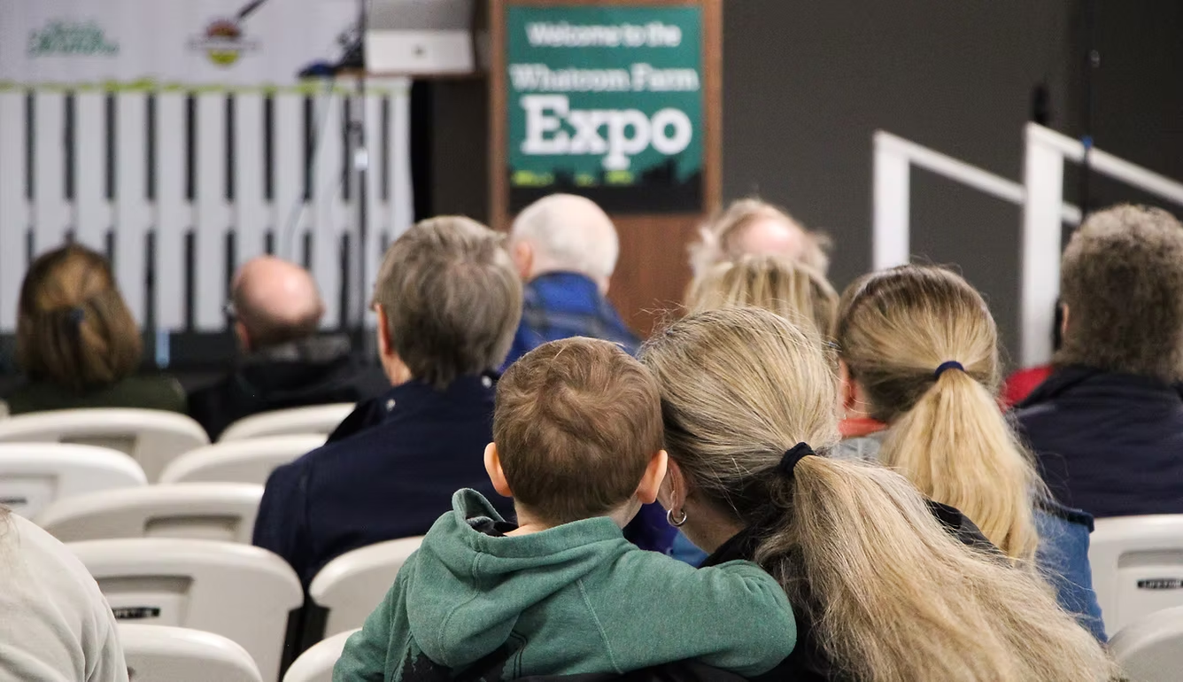 People sitting in chairs at an event; a sign reads "Welcome to the Whatcom Farm Expo."