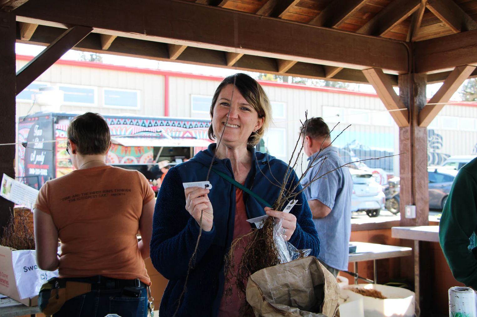 A woman smiling, holding plant saplings and a tag at an outdoor market or event.