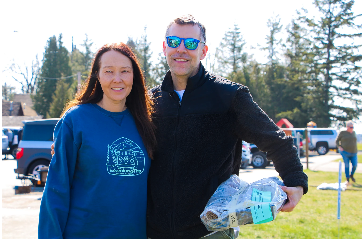 A man and a woman smiling outdoors; he's holding a wrapped item.