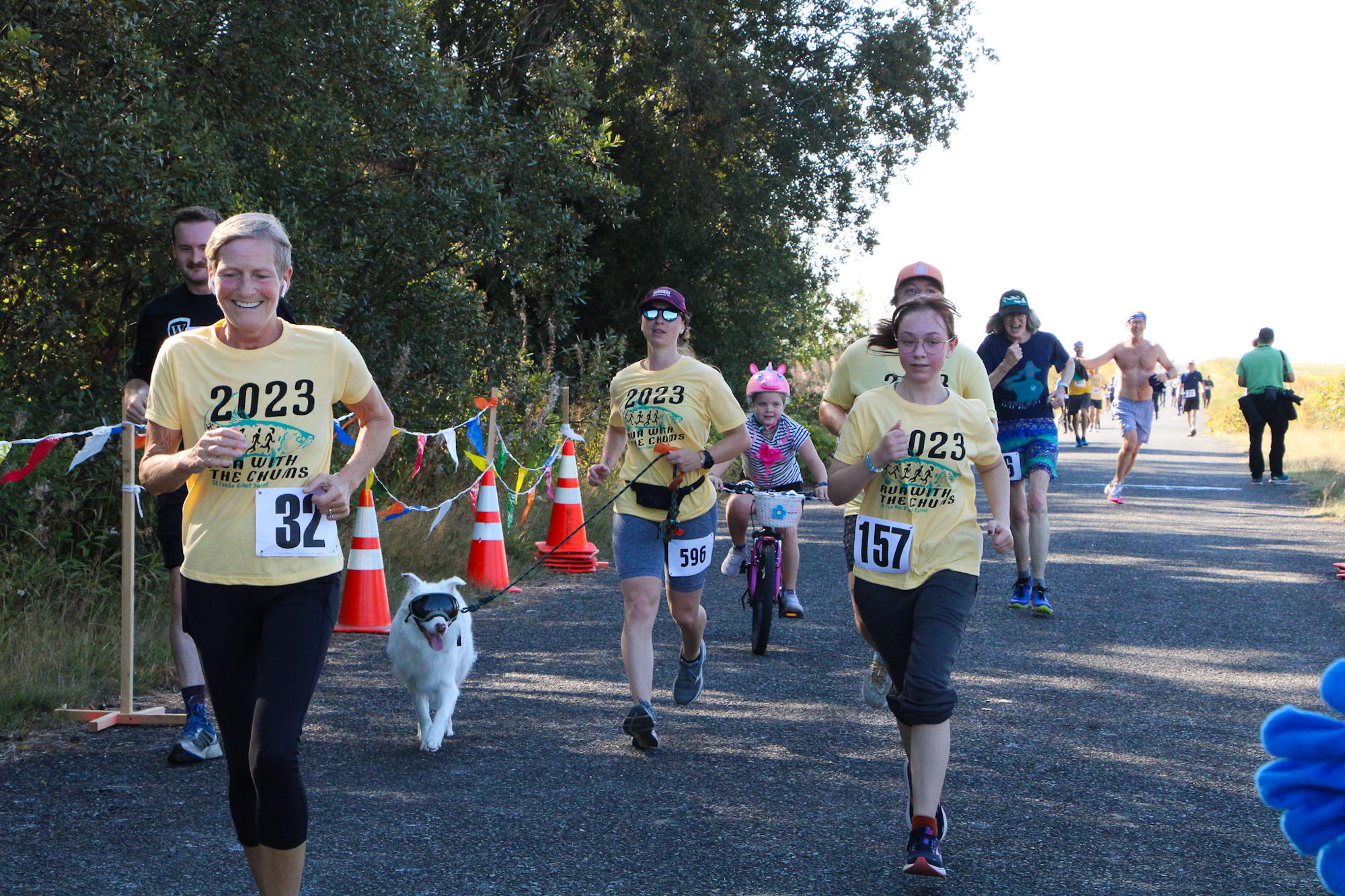 People running in a race, some wearing "2023 Run with the Chums" shirts. A child on a bike and a dog are also participating.