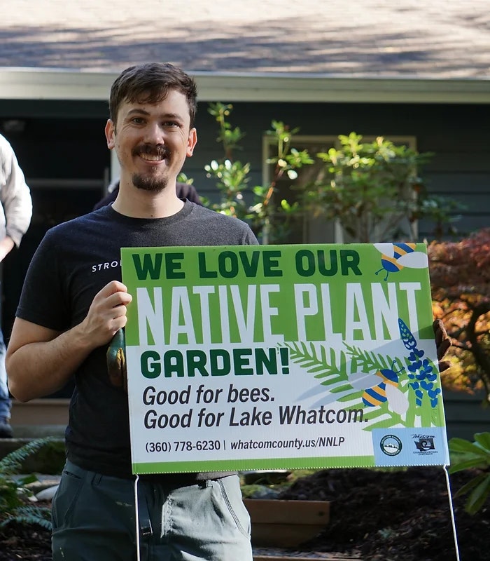 A person holds a sign saying, "We love our native plant garden! Good for bees. Good for Lake Whatcom."