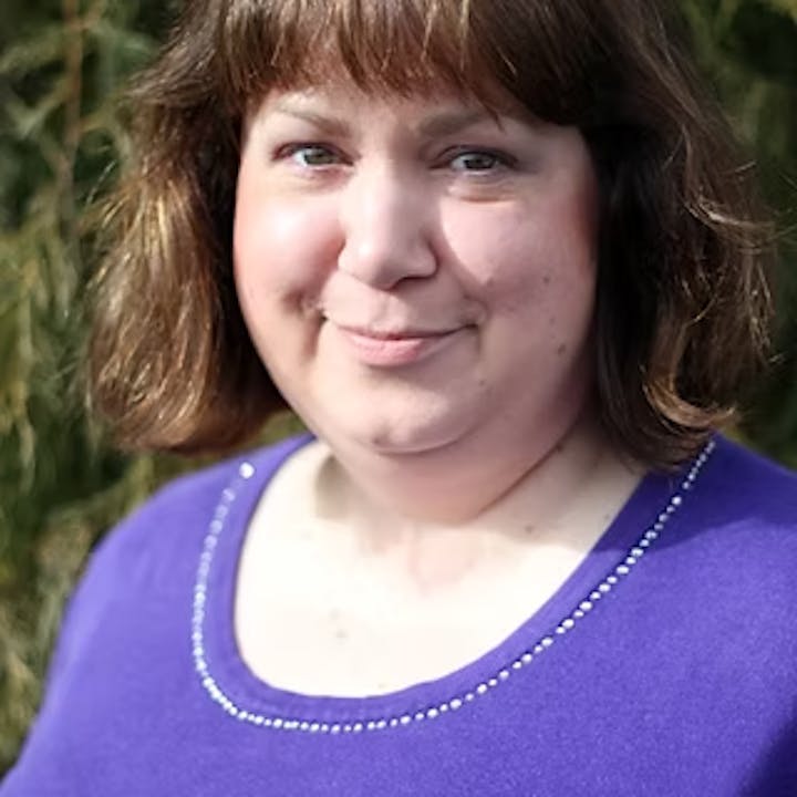 A person with brown hair, wearing a purple top, smiling outside with greenery in the background.