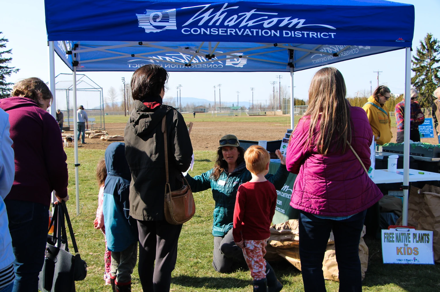 A group of people under a "Whatcom Conservation District" tent, discussing native plants for kids.