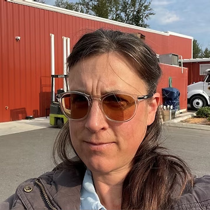 A person wearing sunglasses stands outside a red industrial building on a sunny day.