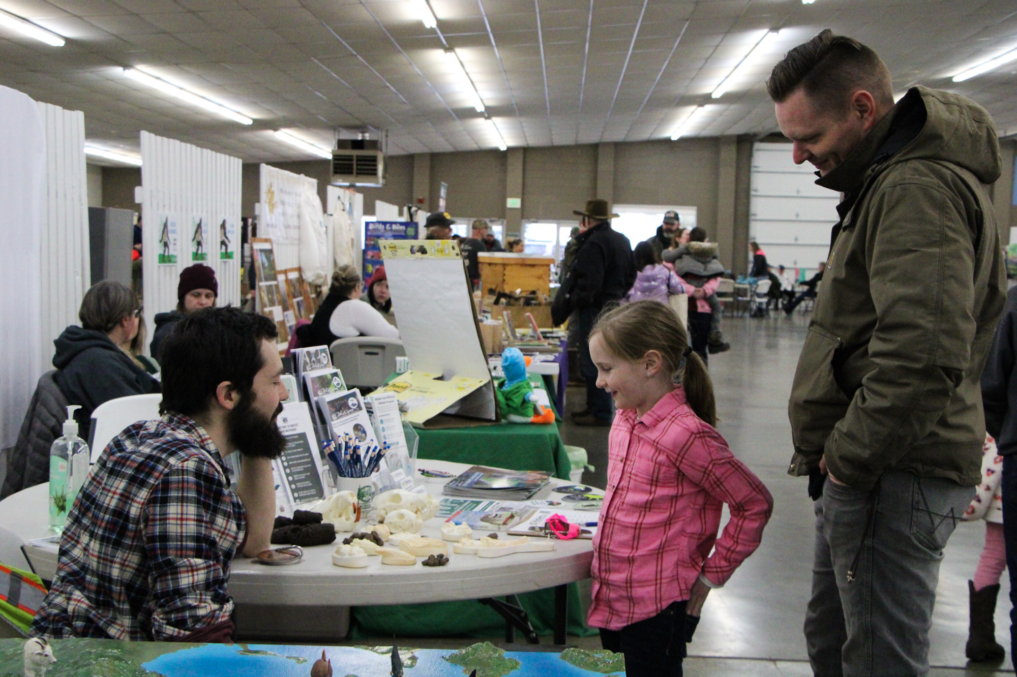 Man and girl at a table with displays in a busy indoor event, people in the background.