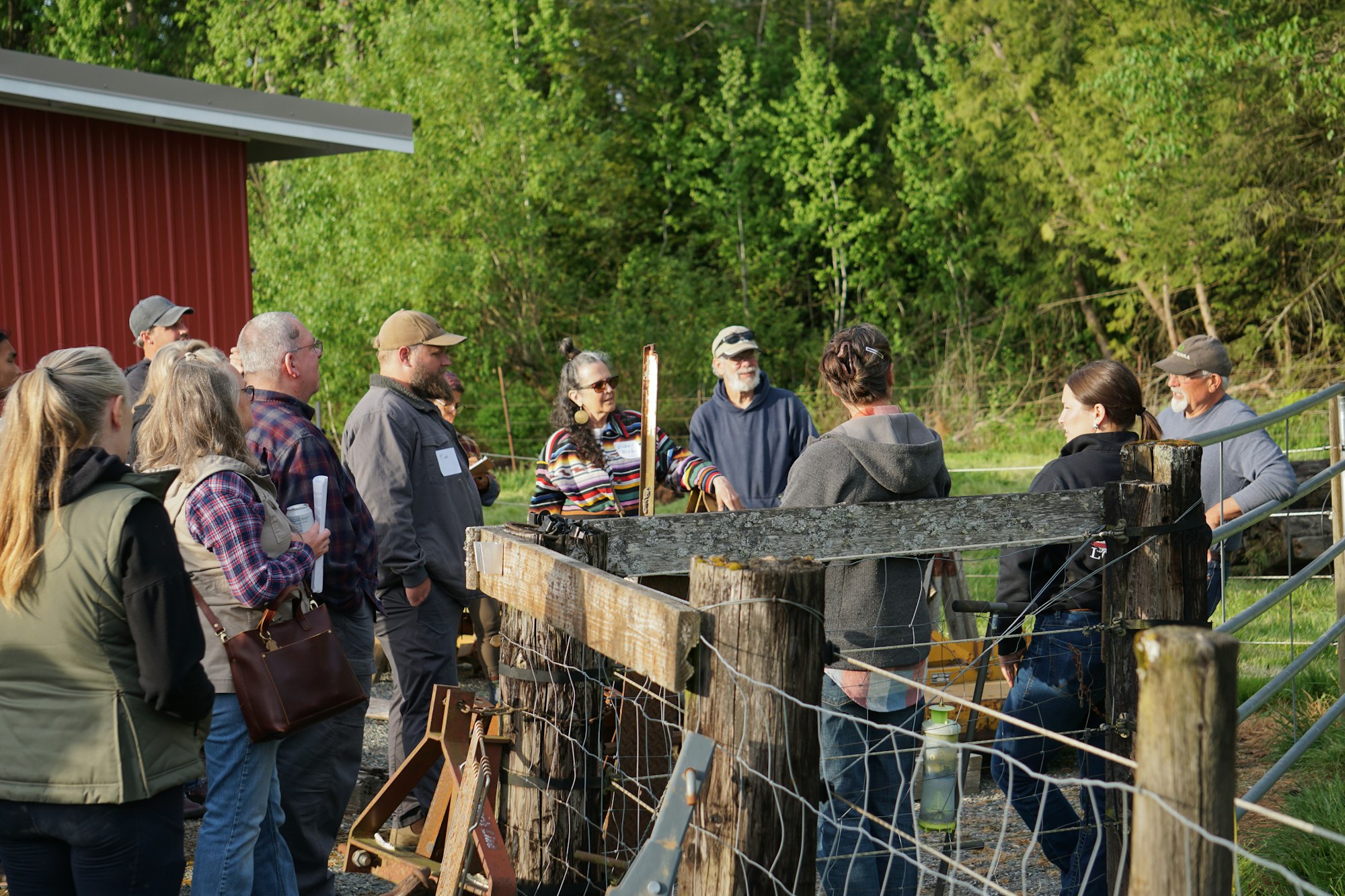 A group of people gathered outside near a red building, discussing something around a fenced area, with trees in the background.