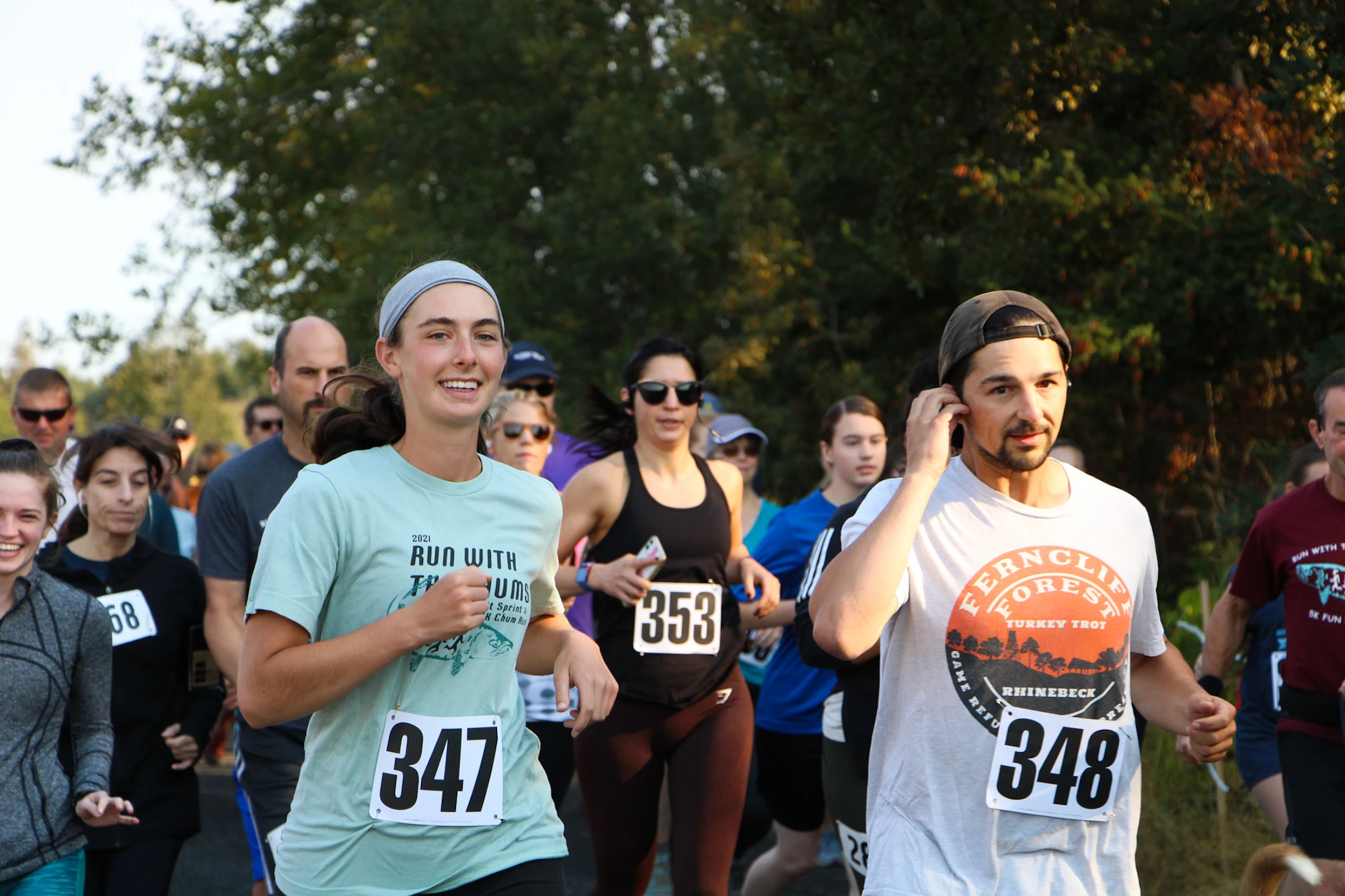 People running in a race, wearing numbered bibs, smiling, with trees in the background.