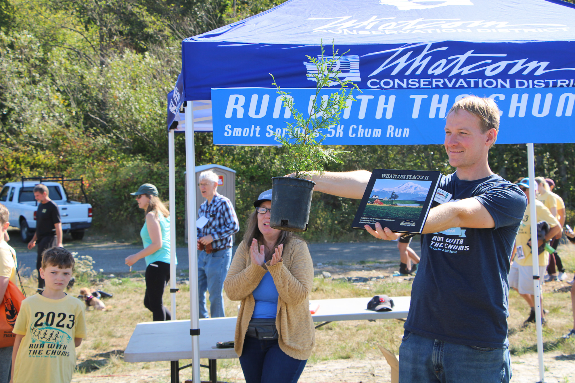 A man holding a potted plant and book at an outdoor event. People and a "Run with the Chums" banner are visible under a tent.