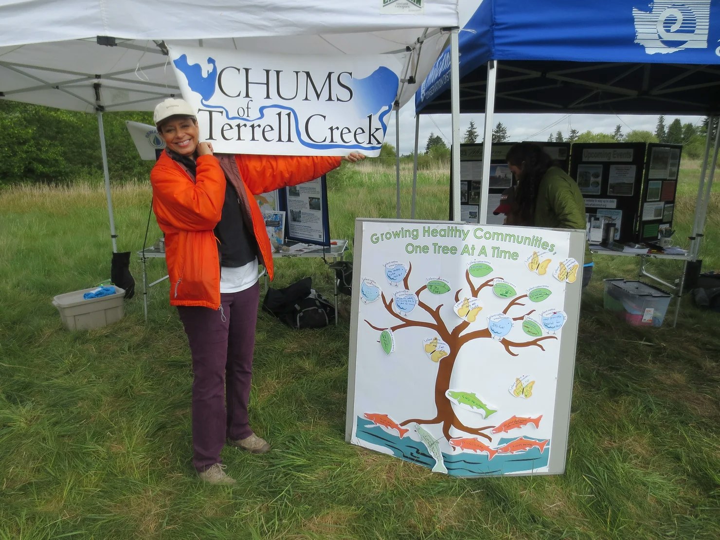 A person holds a "CHUMS of Terrell Creek" sign next to a display board under tents, promoting community growth and environmental care.