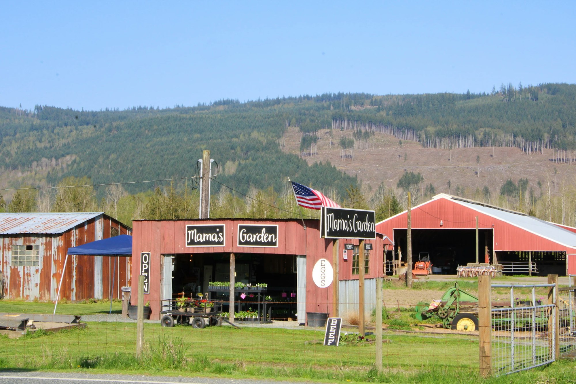 A rural farm stand named "Mama's Garden" with signs for eggs and beef, an American flag, and a barn in a scenic mountainous area.