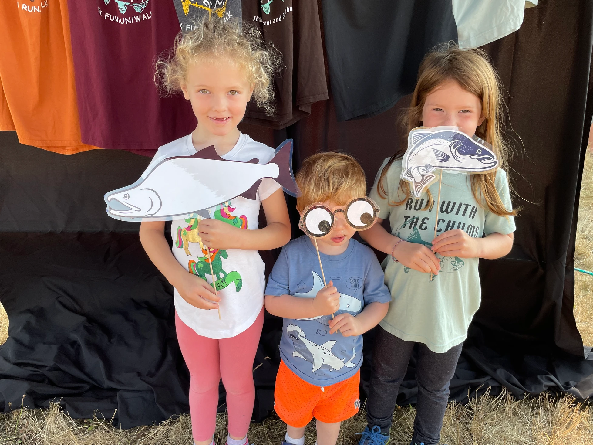 Three children holding fish cutouts and comic eye props, smiling in front of a display with shirts hanging above.