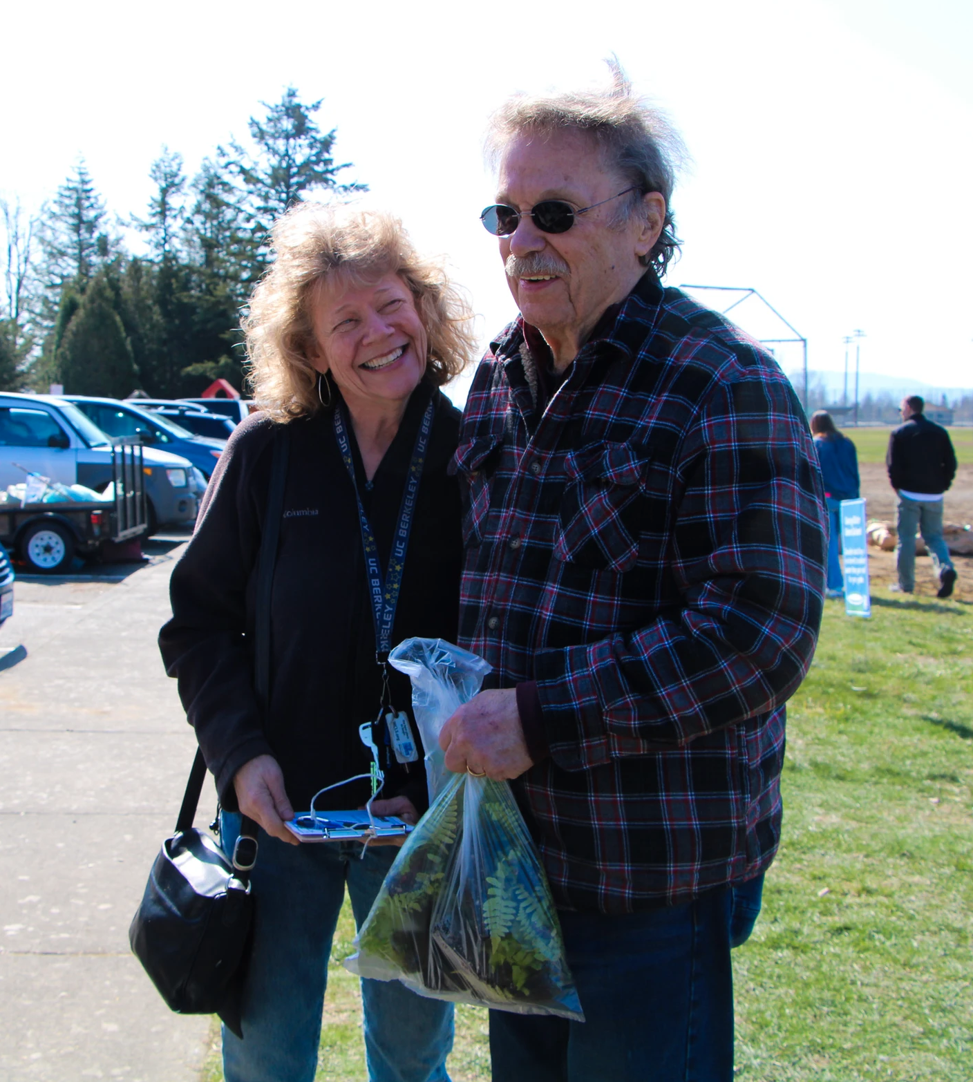 A smiling couple stands outside; the man holds a bag with plants.
