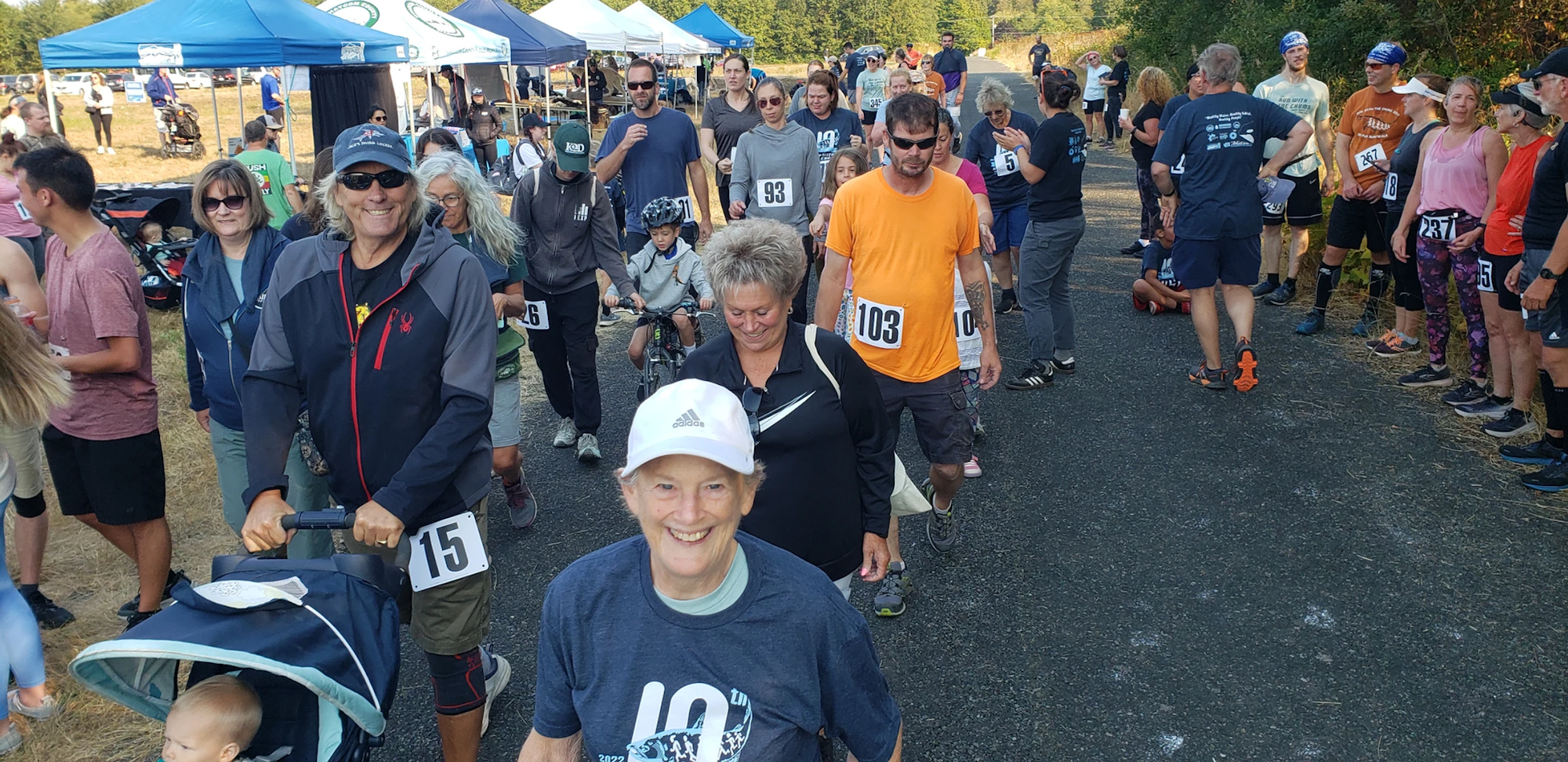 People with number bibs participating in an outdoor event, some pushing strollers and riding bikes, with tents in the background.