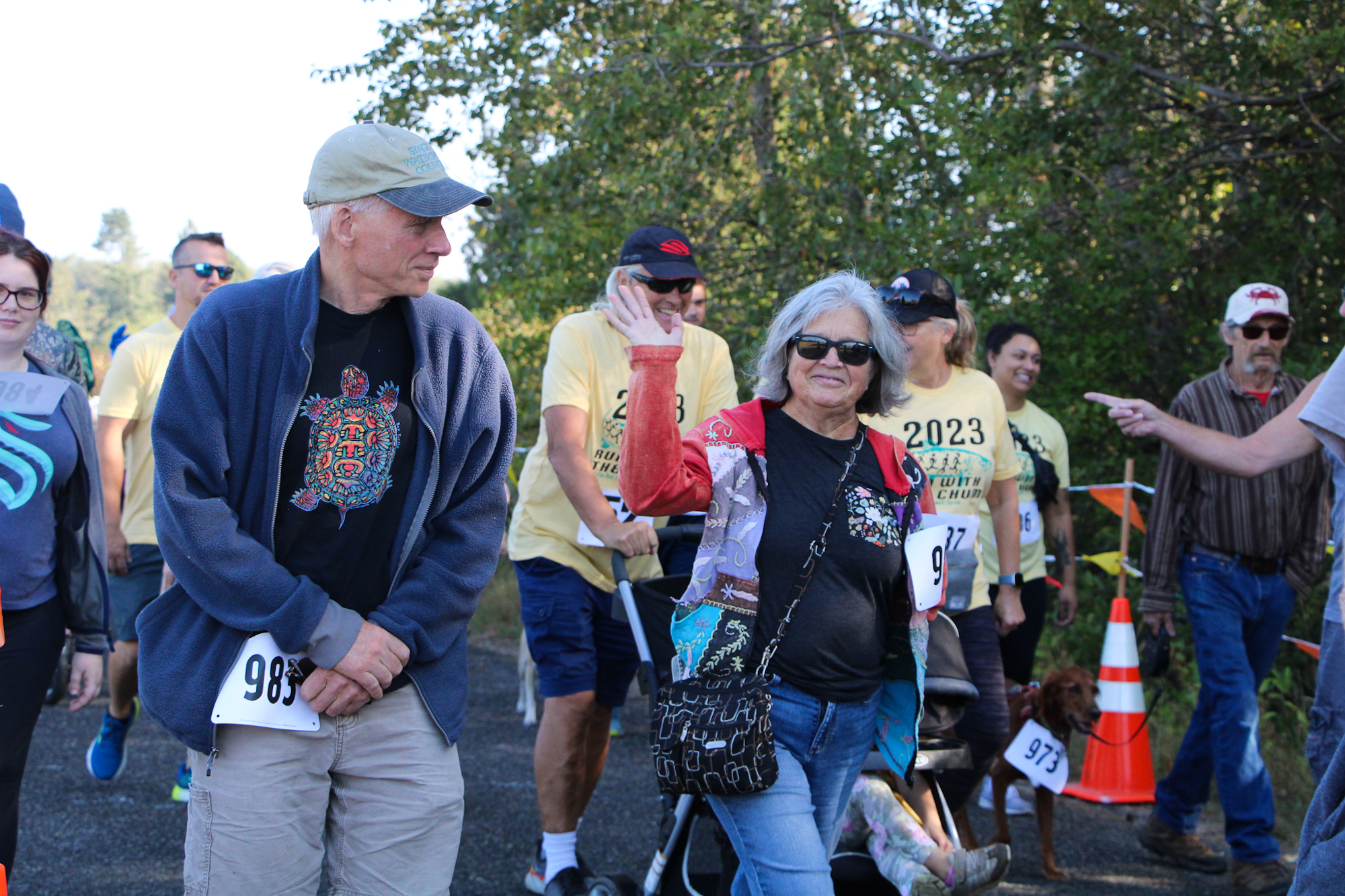 A group of people at an outdoor event, some wearing numbered bibs, walking or jogging. One person is waving.