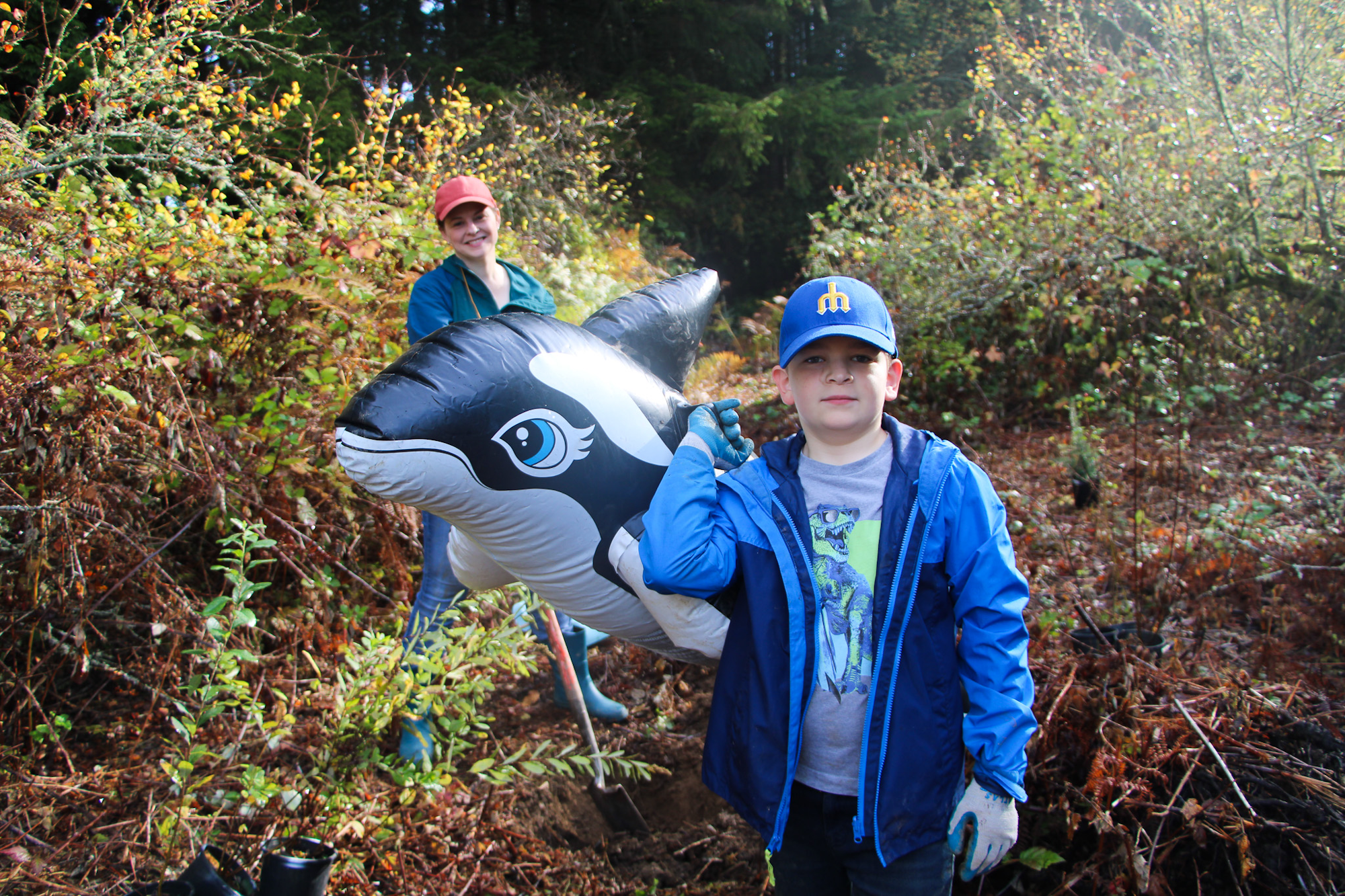 A person and a child outdoors with a large inflatable orca, surrounded by greenery.