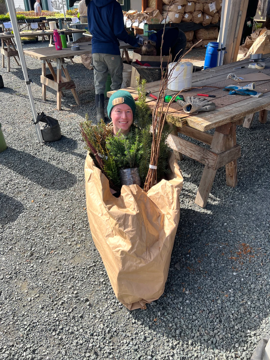 A person smiling inside a large paper bag filled with plants, at a rustic outdoor setting with wooden tables and gardening supplies.