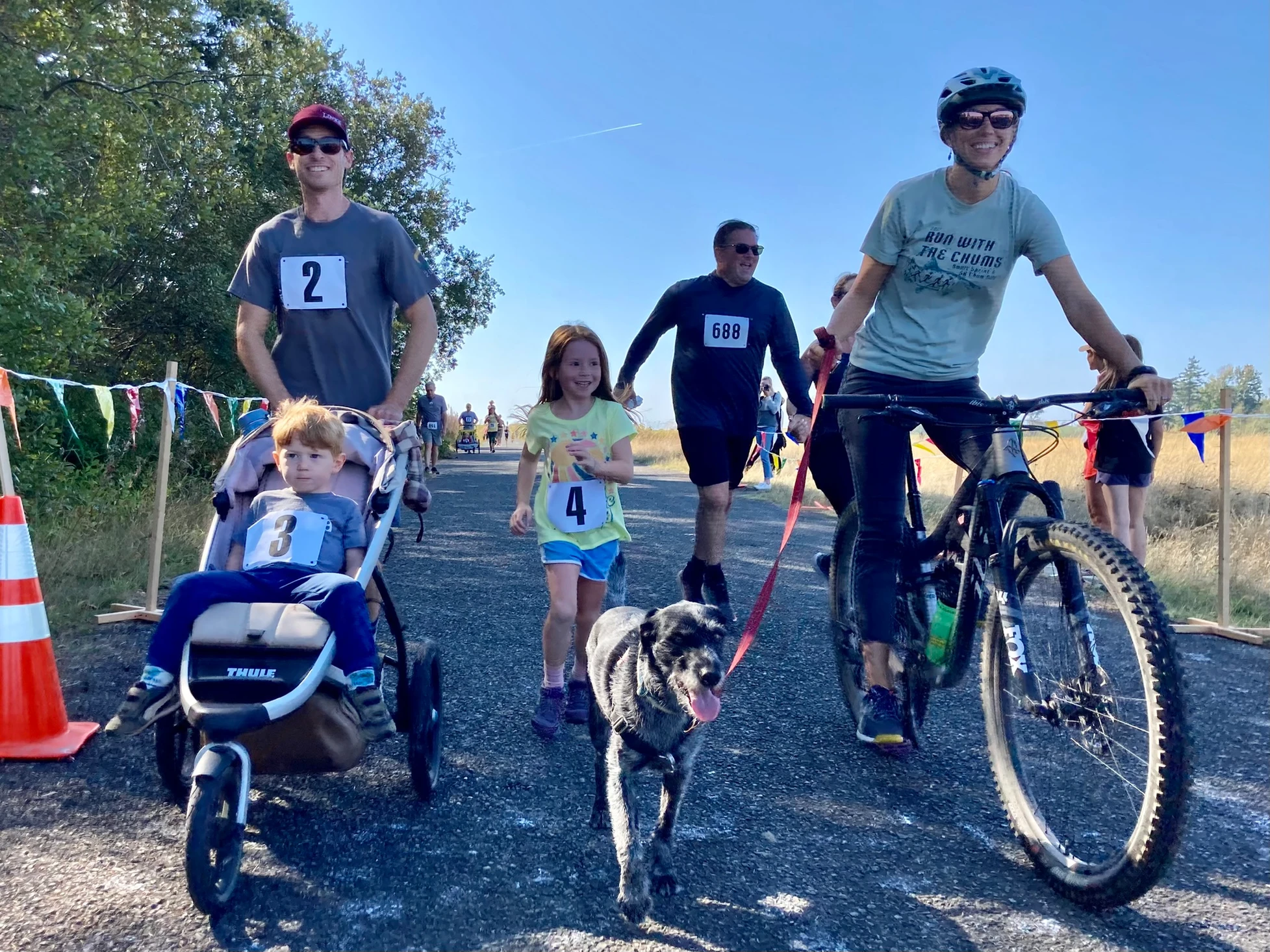 People in a race event. One pushes a stroller, a woman holds a bike, and a man with a dog. They have numbered tags.