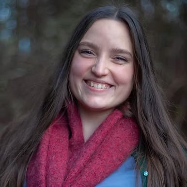 A person with long hair smiling, wearing a red scarf outdoors.