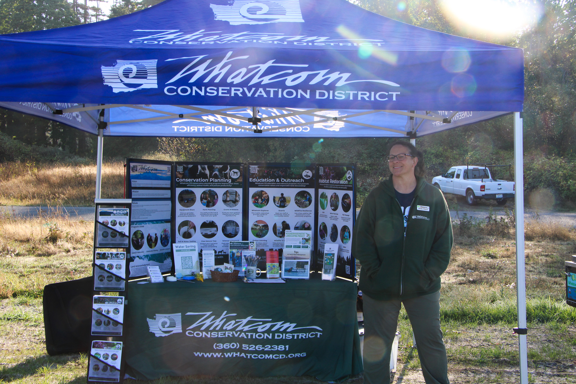 A person stands under a "Whatcom Conservation District" tent with informational displays and brochures.