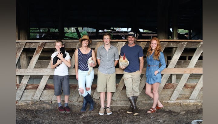 A group of five people, two holding chickens and one holding a black animal, standing in front of a wooden structure.