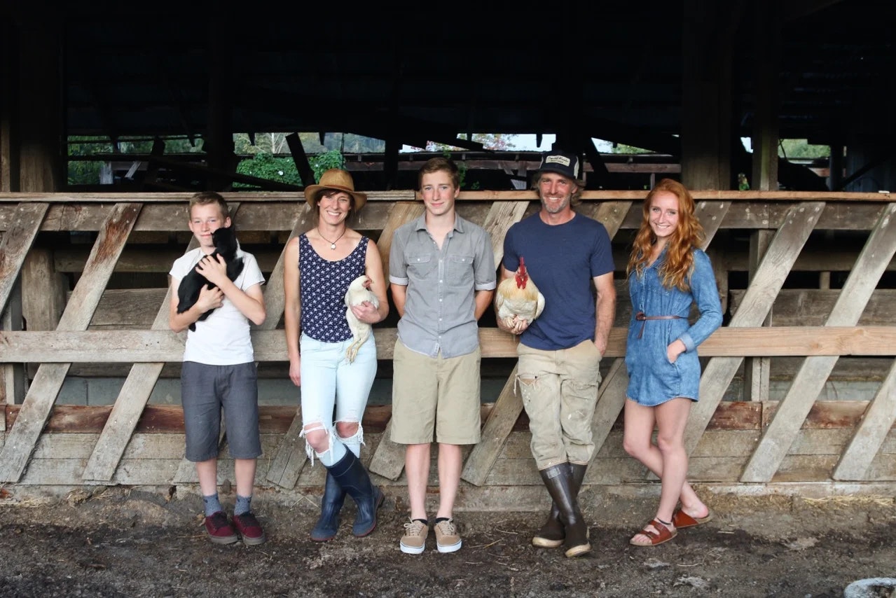 A group of five people, two holding chickens and one holding a black animal, standing in front of a wooden structure.
