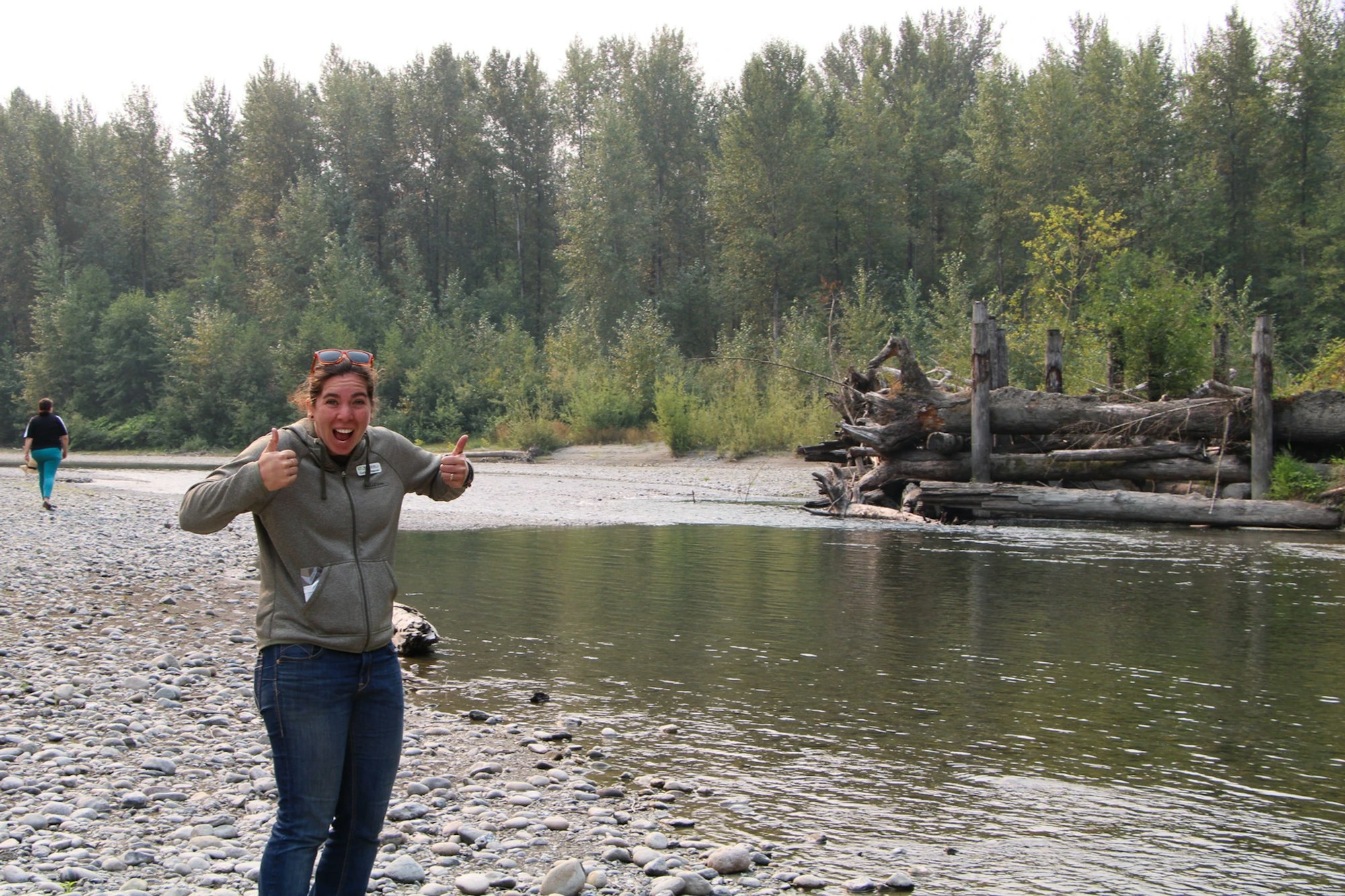 Person giving thumbs up by a river, with trees and logs in the background. Another person walks away in the distance.