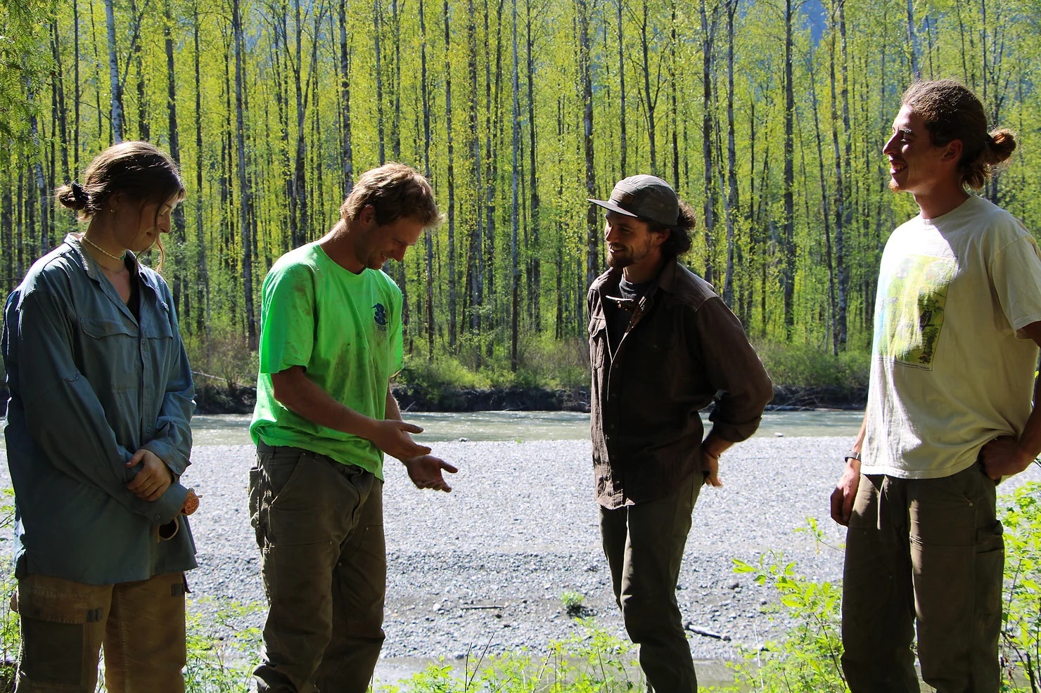 Four people standing and talking near a river with tall trees in the background.