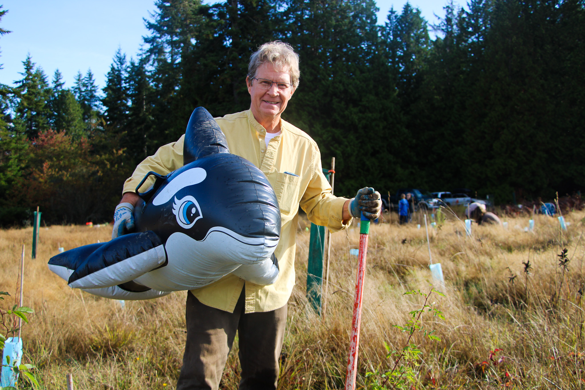 A person holding an inflatable orca stands in a field with planting tools, surrounded by trees.