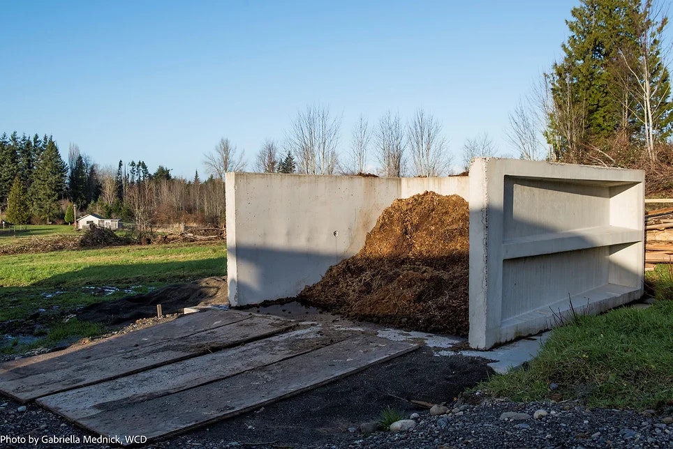 A concrete manure storage structure partially filled, surrounded by grass and trees under a clear sky.