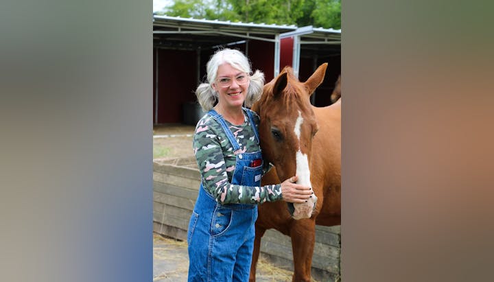 A person wearing overalls and a camo shirt stands beside a brown horse, smiling outdoors.