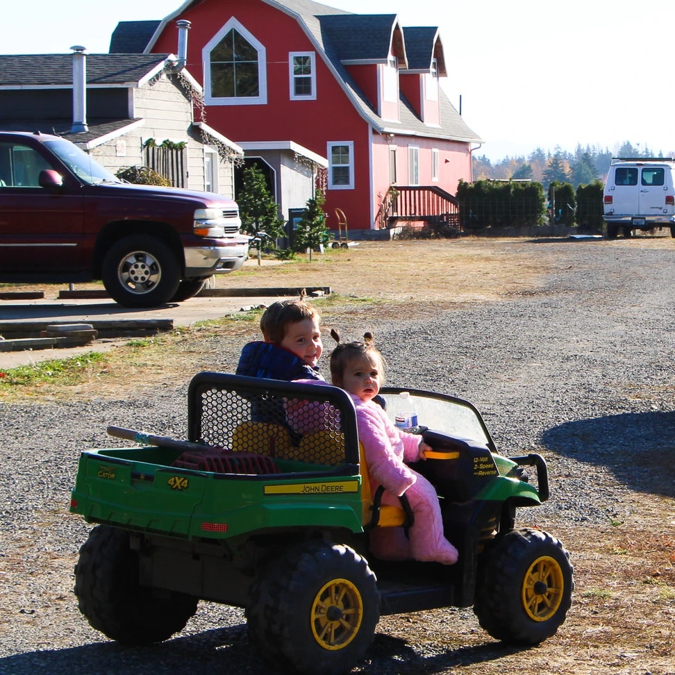 Two kids riding a toy John Deere vehicle in front of a red house.