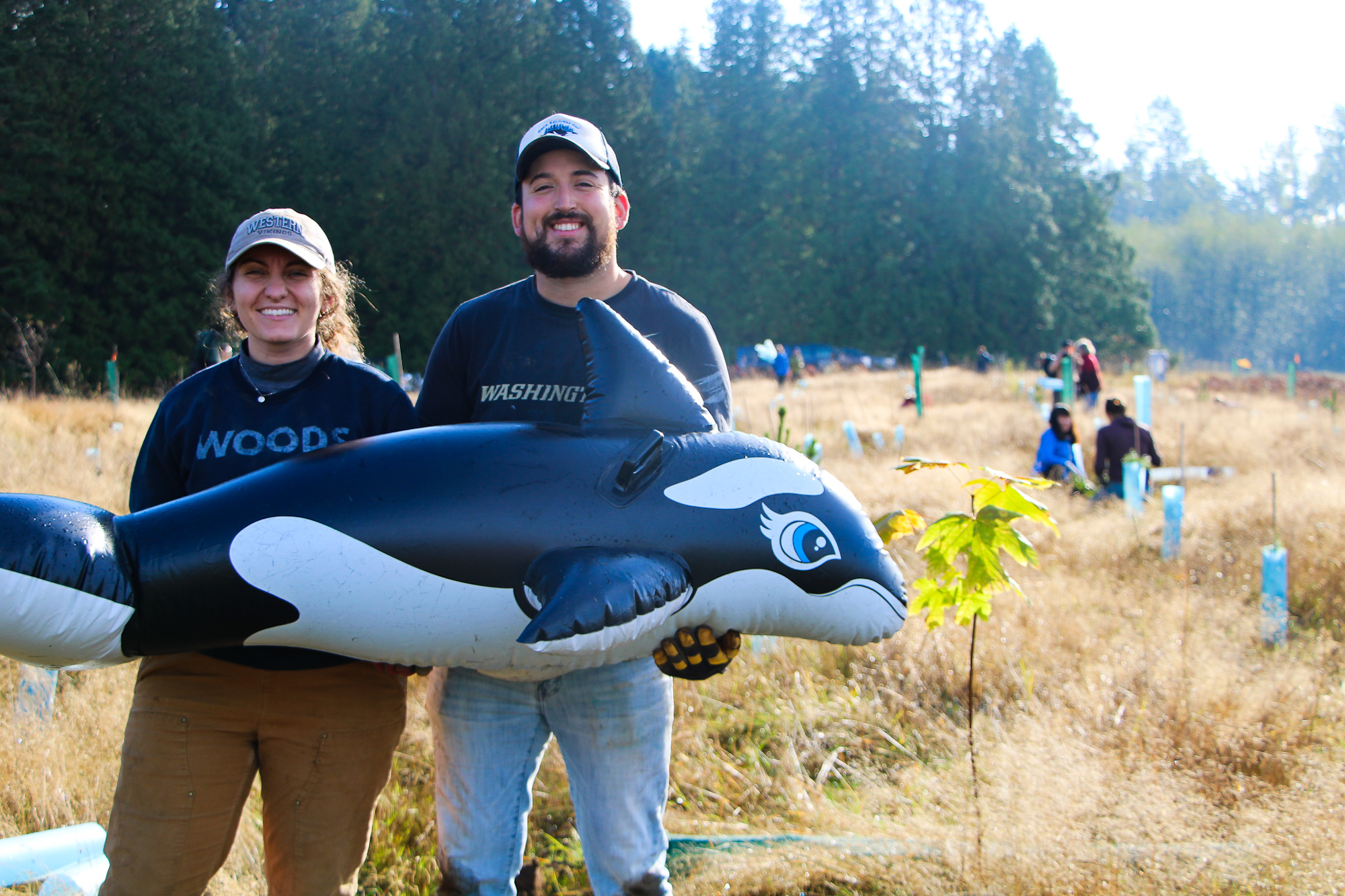 Two people outdoors holding an inflatable orca amidst a field with young trees and people in the background.
