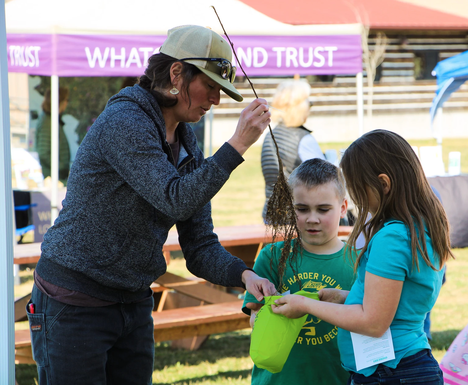 A woman shows two children a plant root at an outdoor event.