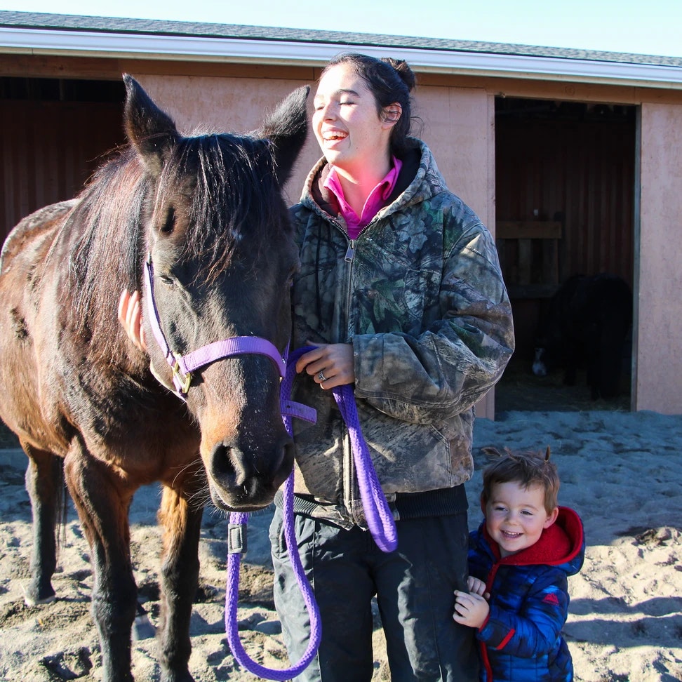 A person and a child smiling with a horse in a stable area.