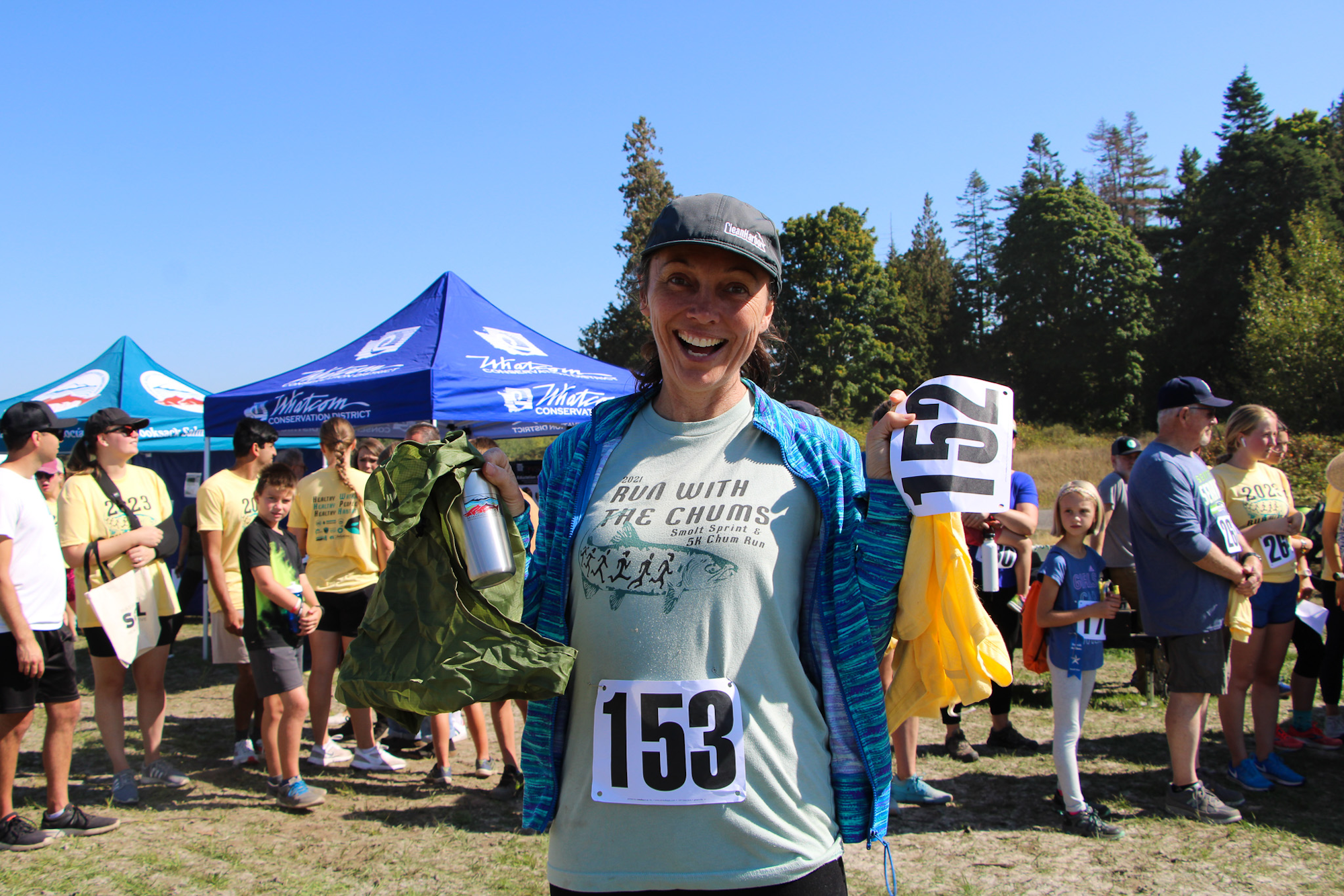 A woman smiling with race bib number 153, standing in a crowd at an outdoor event.