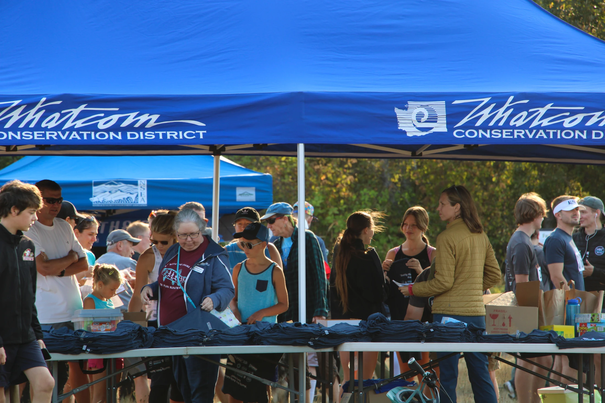 People gathered under a "Whatcom Conservation District" tent, distributing items at an outdoor event.